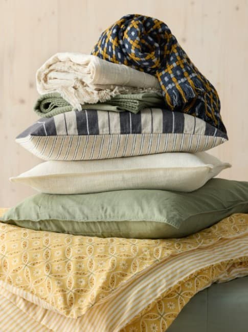 Pila de almohadas y mantas dobladas con estampados en verde, amarillo y azul sobre una cama con ropa de cama estampada en amarillo.