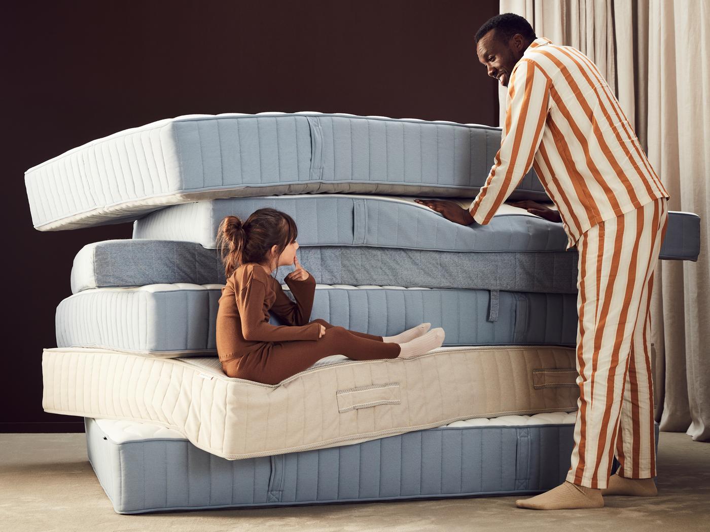 Pile de matelas sur laquelle s’appuie un homme. Une enfant est assise sur l’un des matelas.