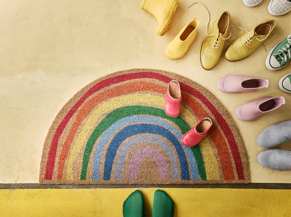 PILLEMARK door mat with a rainbow pattern lying on a yellow floor with colourful shoes and a pair of feet with green socks.