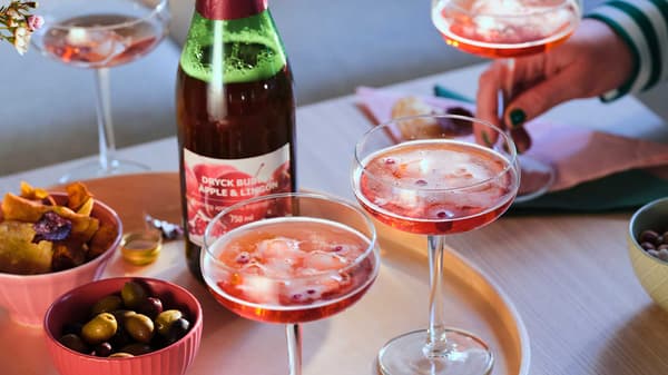 Pink sparkling beverage in tall-stemmed glasses on a tray with small bowls of olives and chips.