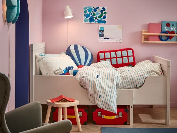 ピンクの壁に白いベッドを置いた子ども部屋。赤い収納とカラフルな寝具で明るい雰囲気。