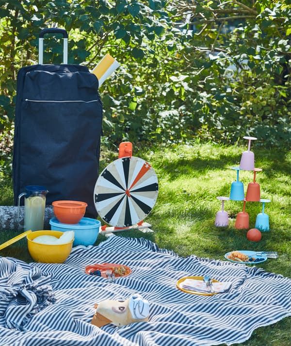 Pique-nique sur l’herbe avec vaisselle, jeux et une grande couverture, transportés dans un grand sac de sport à roulettes bleu FÖRENKLA.