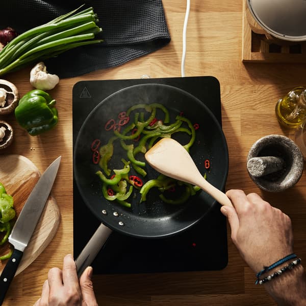 Piros és zöld paprikát kevernek egy serpenyőben, egy hordozható IKEA TILLREDA indukciós főzőlapon, egy konyhapulton.