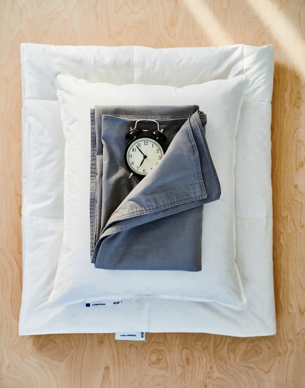 Piumino bianco piegato, cuscino bianco, lenzuolo blu scuro e una sveglia nera dallo stile vintage, su una superficie in legno.