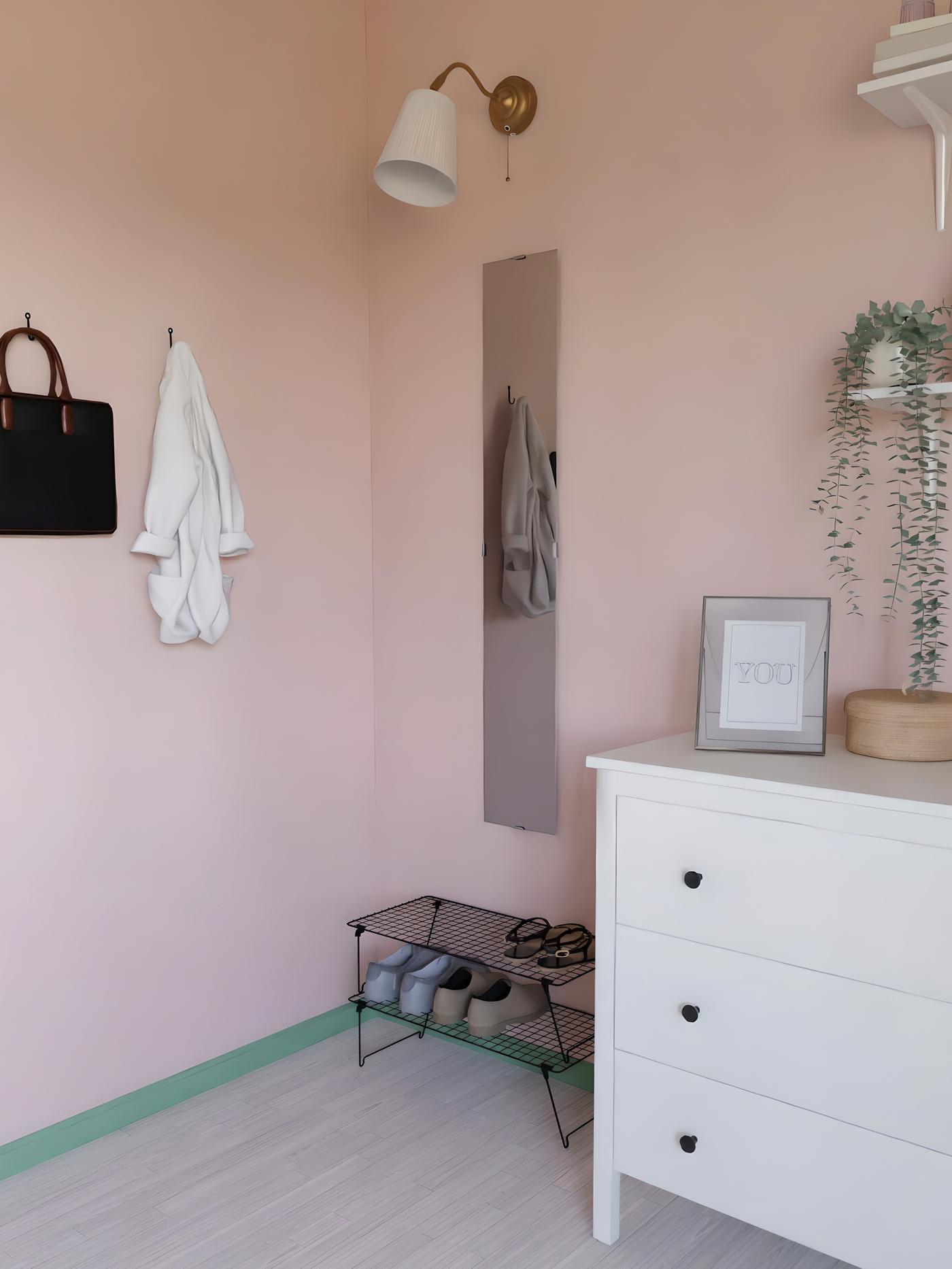 Pla d’una habitació rosa amb una còmoda, un moble per a sabates, un mirall estret i penjadors amb roba.