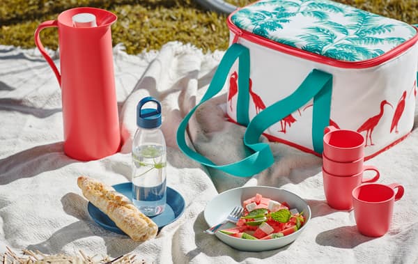 Plaid blanc installé dans l’herbe pour un pique-nique, sur lequel se trouvent un sac isotherme SOMMARLIV, des mugs TALRIKA et une salade d’été servie dans une assiette TALRIKA.