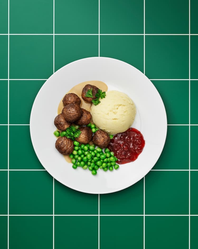 Plain white plate of plant-based meatballs, brown sauce, mash potatoes, lingonberry jam, green peas and a sprig of parsley.