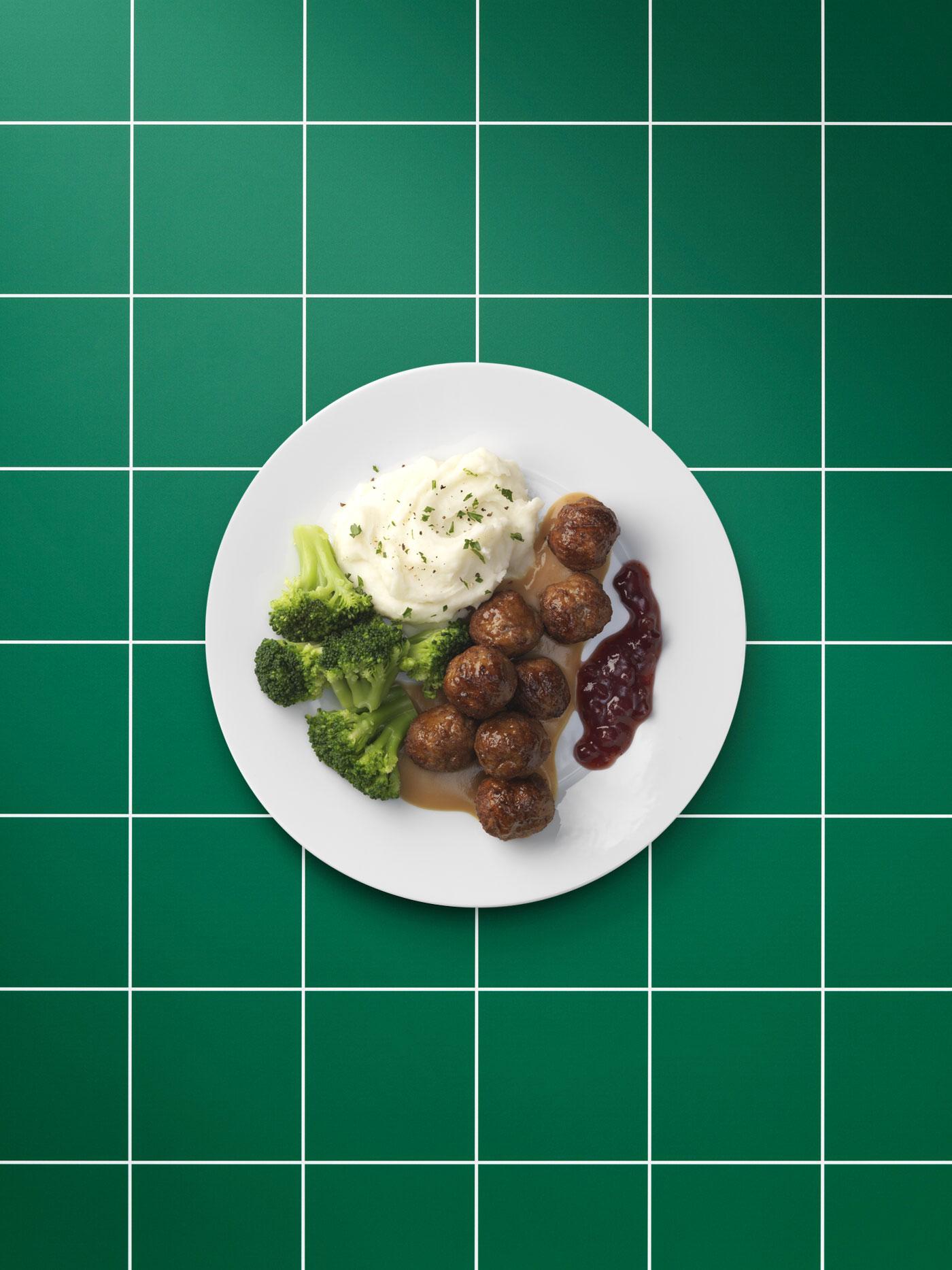 Plain white plate of plant-based meatballs, brown sauce, mashed potatoes, lingonberry jam, brocolli and a sprig of parsley.