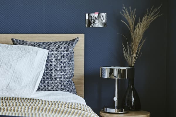 Plan rapproché sur le coin d’un lit, une table de chevet et une lampe contre un mur bleu foncé.