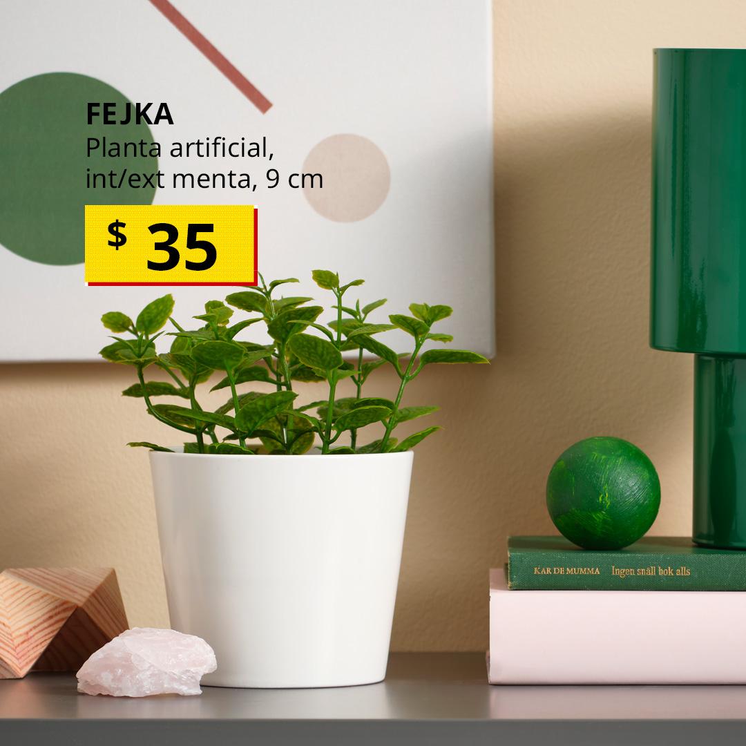 Planta artificial FEJKA tipo menta para interior y exterior en una maceta blanca sobre un mueble junto a varios artículos de decoración.