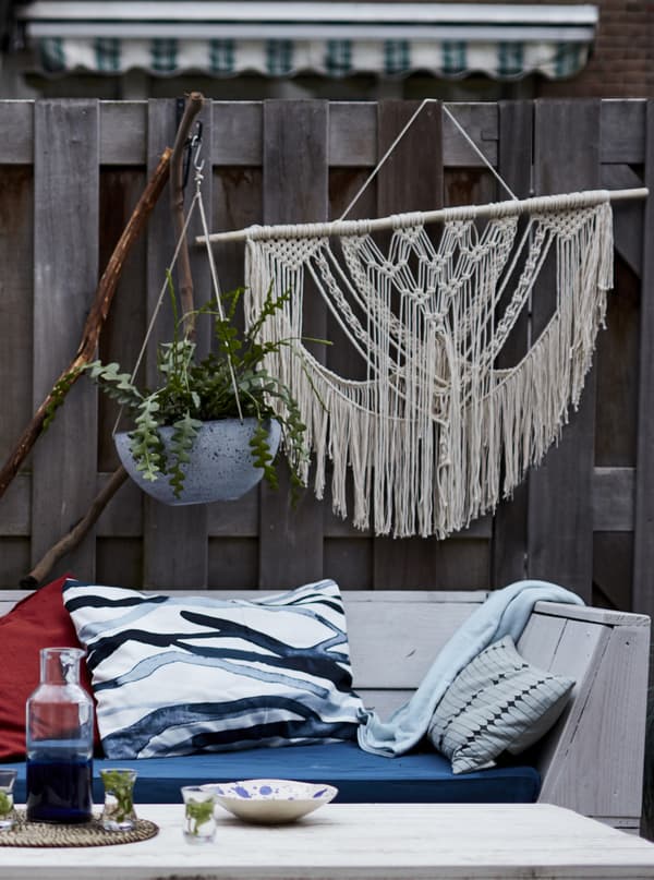 Plants and macramé hanging on a fence behind a wooden bench with cushions.