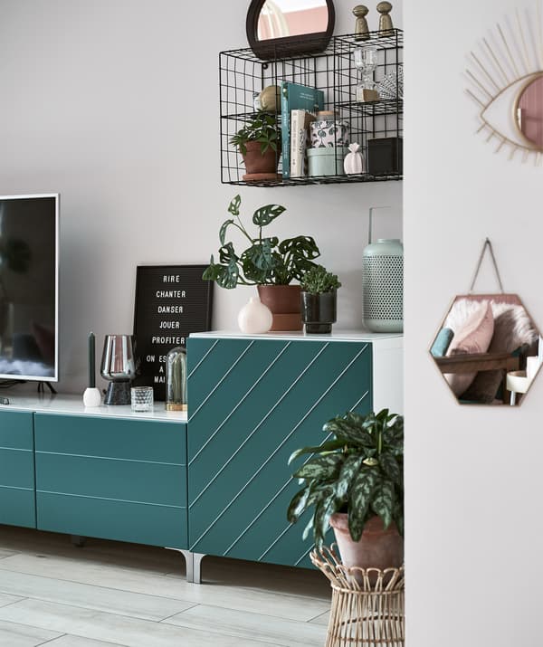 Plants and ornaments on a low TV unit with green doors and a black wire shelving unit on the wall.