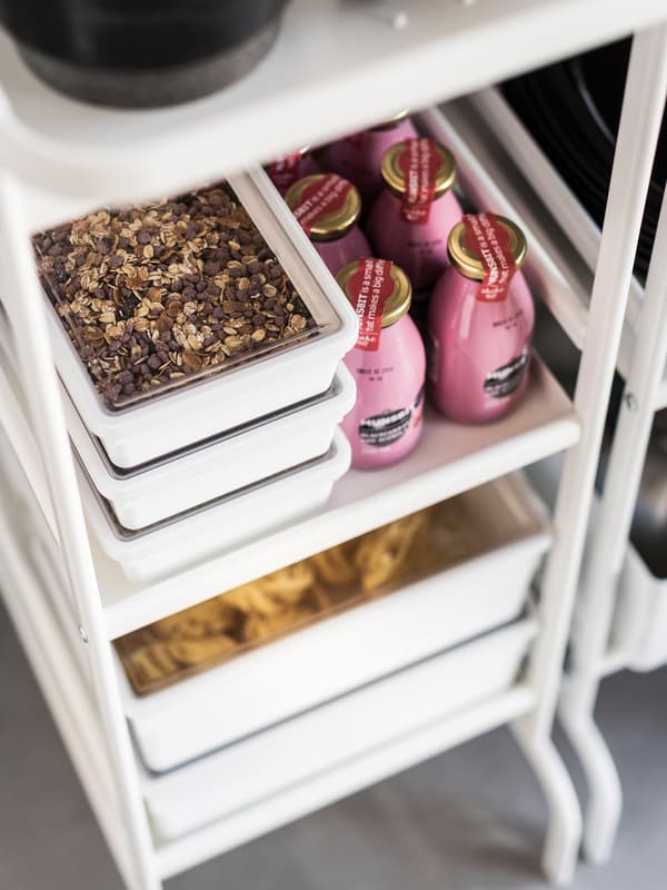 Plastic containers containing muesli with see-through lids on a white trolley next to pink bottles of smoothies.