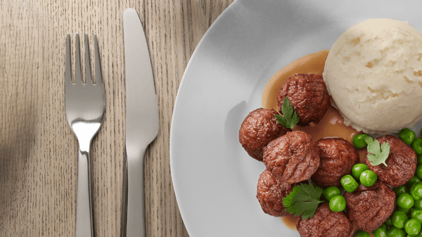 Plate with meatballs in gravy, mashed potatoes, and green peas, with fork and knife on a wooden table.
