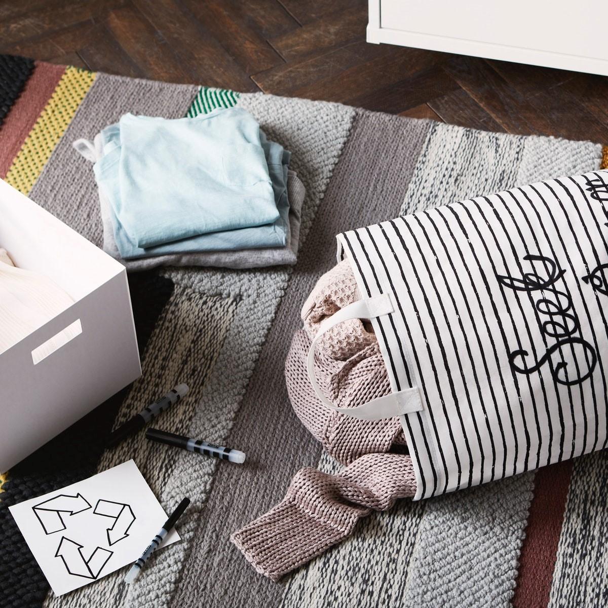 Plátené vrecko s nápisom „Hľadá sa nový majiteľ“ s oblečením vo vnútri na koberci vedľa škatule, ceruziek a symbolu recyklácie.