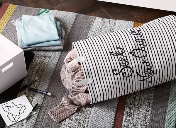 Plátené vrecko s nápisom „hľadá sa nový majiteľ“ s oblečením vo vnútri na koberci vedľa škatule, ceruziek a symbolu recyklácie.