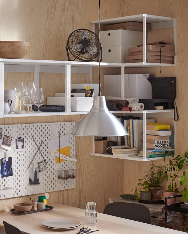 PLATSA white shelving units are wall-mounted in a corner. Books, glasses, a mini fan and more are stored on the shelves.