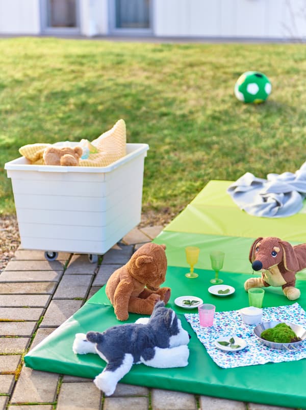 Playful dinner for a circle of soft-toys animals on a PLUFSIG mat by a patch of grass. A SOCKERBIT box with more toys nearby.