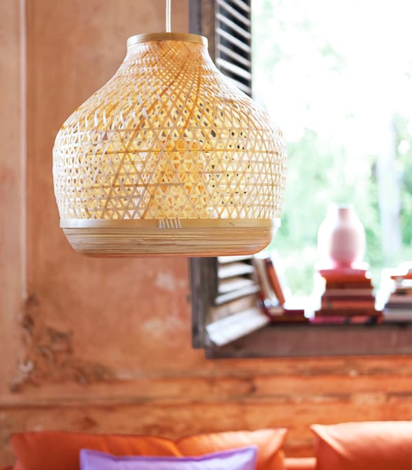 Pletená závesná lampa pred oranžovou stenou so žalúziami z tmavého dreva a knihami v pozadí.