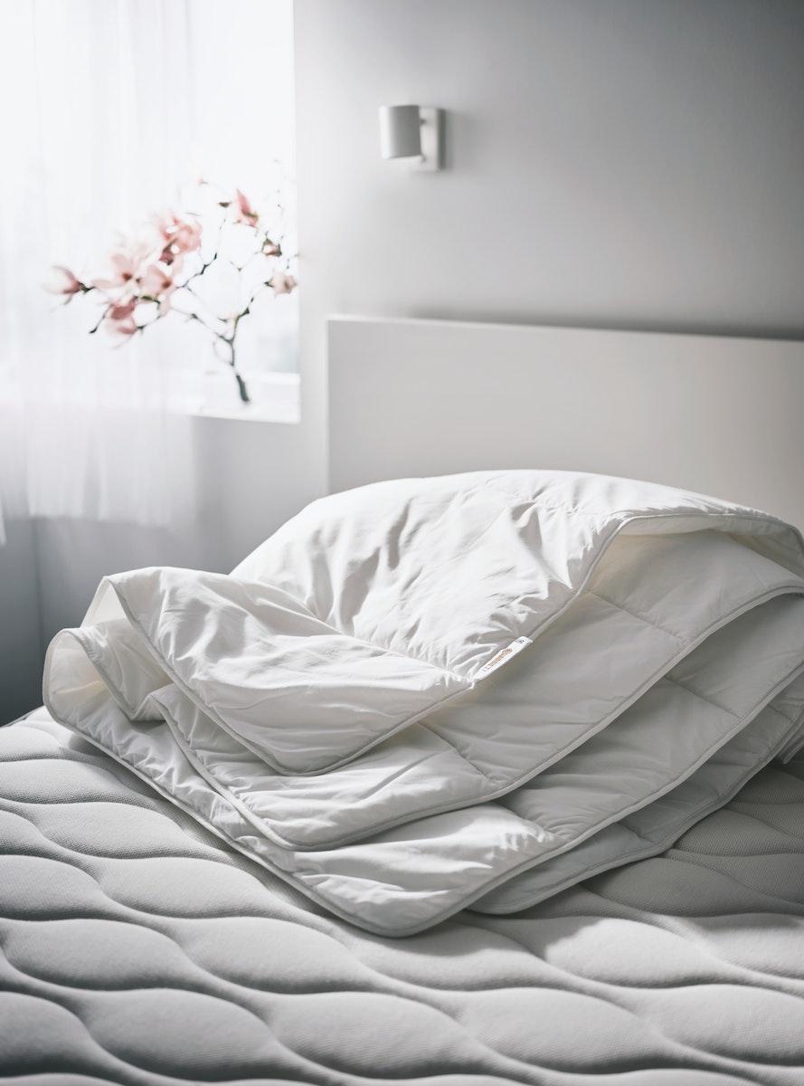 Plumón SMÅSPORRE blanco sobre una cama IKEA. La cama no tiene sábanas y se ve el diseño del colchón. La cama tiene un respaldo blanco y de fondo se observa la muralla y un pedazo de ventana cubierto por cortinas blancas.