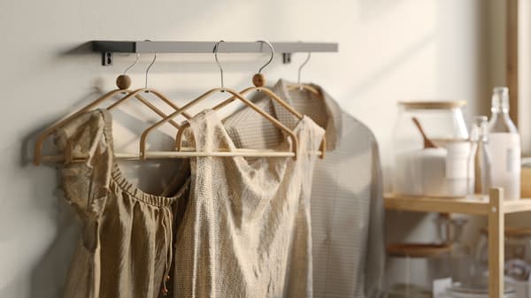 Plusieurs cintres HÖSVANS en bambou sur une barre murale en métal où sont suspendues des chemises et des robes beiges sèches à côté d’une table.