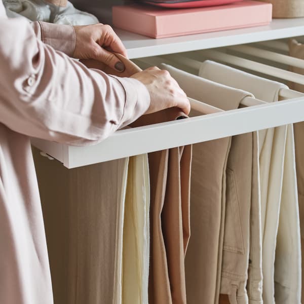 Plusieurs pantalons beiges sur un porte-pantalons coulissant KOMPLEMENT. Une personne en chemisier rose repositionne l’un d’entre eux.