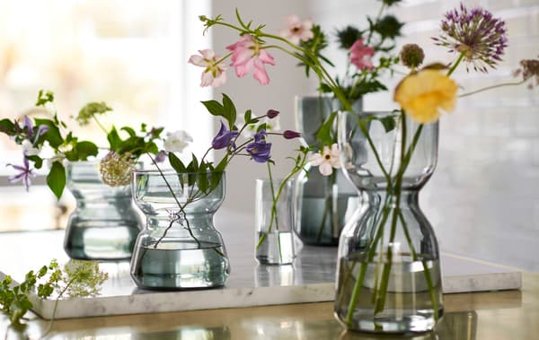 Plusieurs vases OMTÄNKSAM contenant des fleurs, plus étroits sur le milieu pour faciliter la prise en main, sont posés sur un plan de travail.