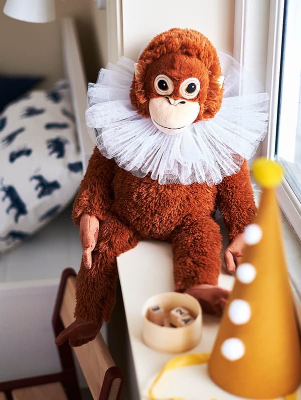 Plyšová opica DJUNGELSKOG sedí na okennom parapete, na krku má biely golier a pred sebou žltú šašovskú čapicu.
