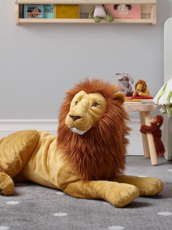 Plyšový lev DJUNGELSKOG leží na sivom koberci, za ním sú malé plyšové zvieratká DJUNGELSKOG na stoličke FLISAT.