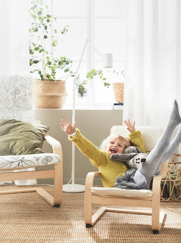 เด็กผู้หญิงนอนเหยียดแขนเหยียดขากับตุ๊กตาผ้าอย่างมีความสุขในอาร์มแชร์ POÄNG/พัวแอง สำหรับเด็กซึ่งตั้งอยู่ถัดจากอาร์มแชร์ขนาดปกติ