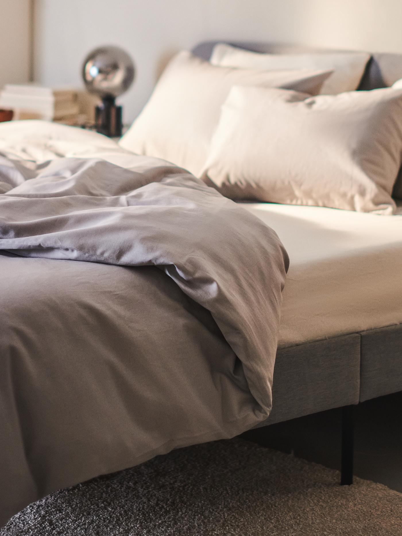 Pod ścianą stoi łóżko tapicerowane SLATTUM z jasnoszarą ramą i materacem piankowym ÅKREHAMN, na którym leży pościel NATTSVÄRMARE.