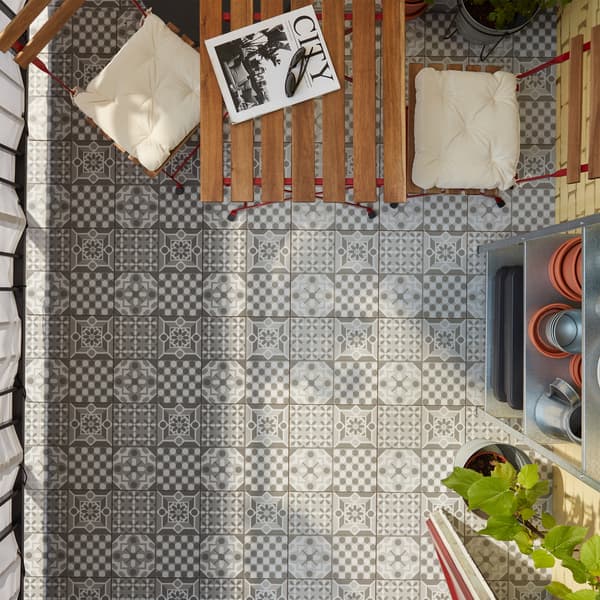 Podeaua de exterior MÄLLSTEN cu modele gri/albe, o masă și scaune din lemn roșu/maro deschis băițuit și o etajeră.