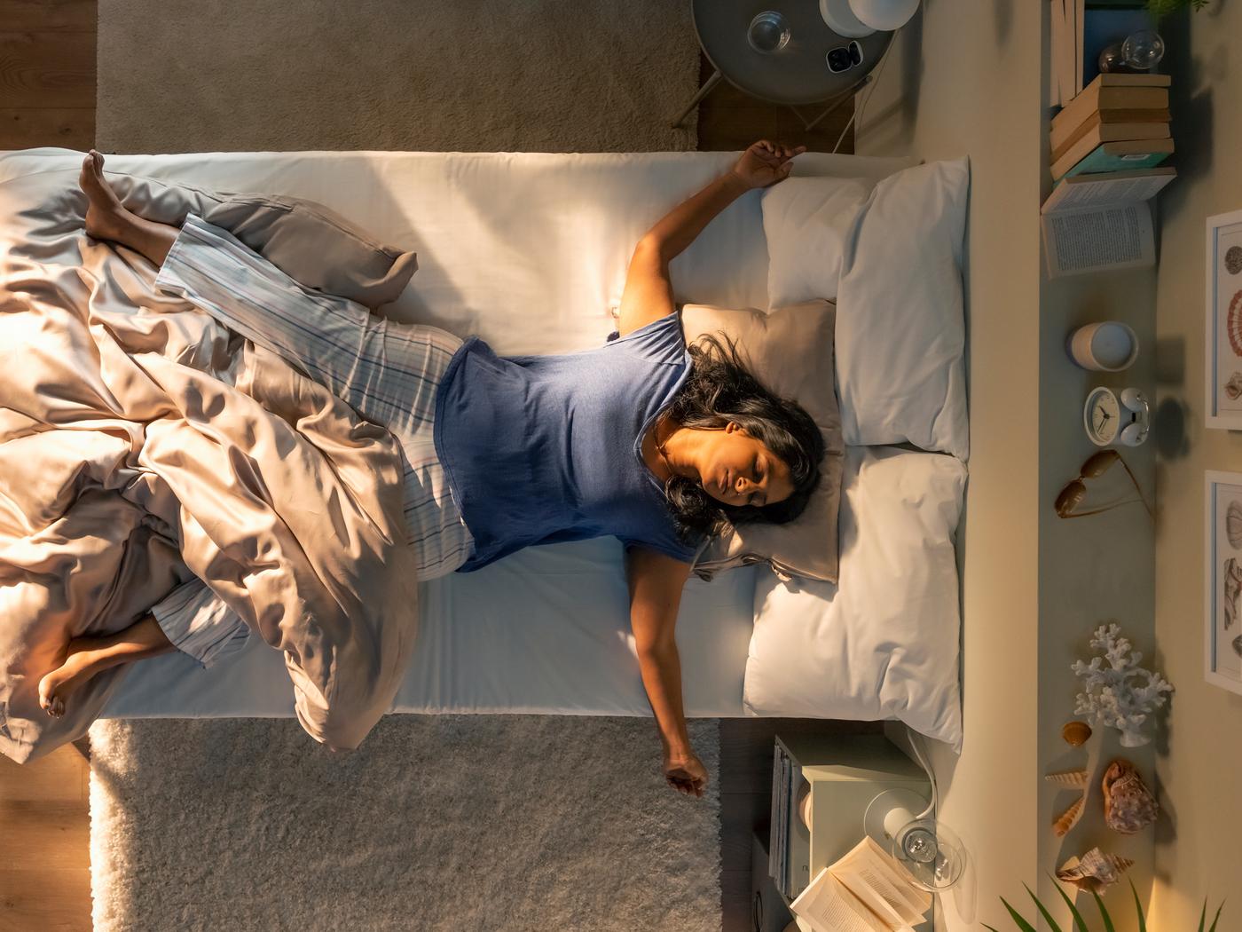 Pogled od zgoraj na osebo, ki spi na hrbtu na postelji, delno pokrita z bež KRITSUGA prevleko za prešito odejo z vzglavnikom.