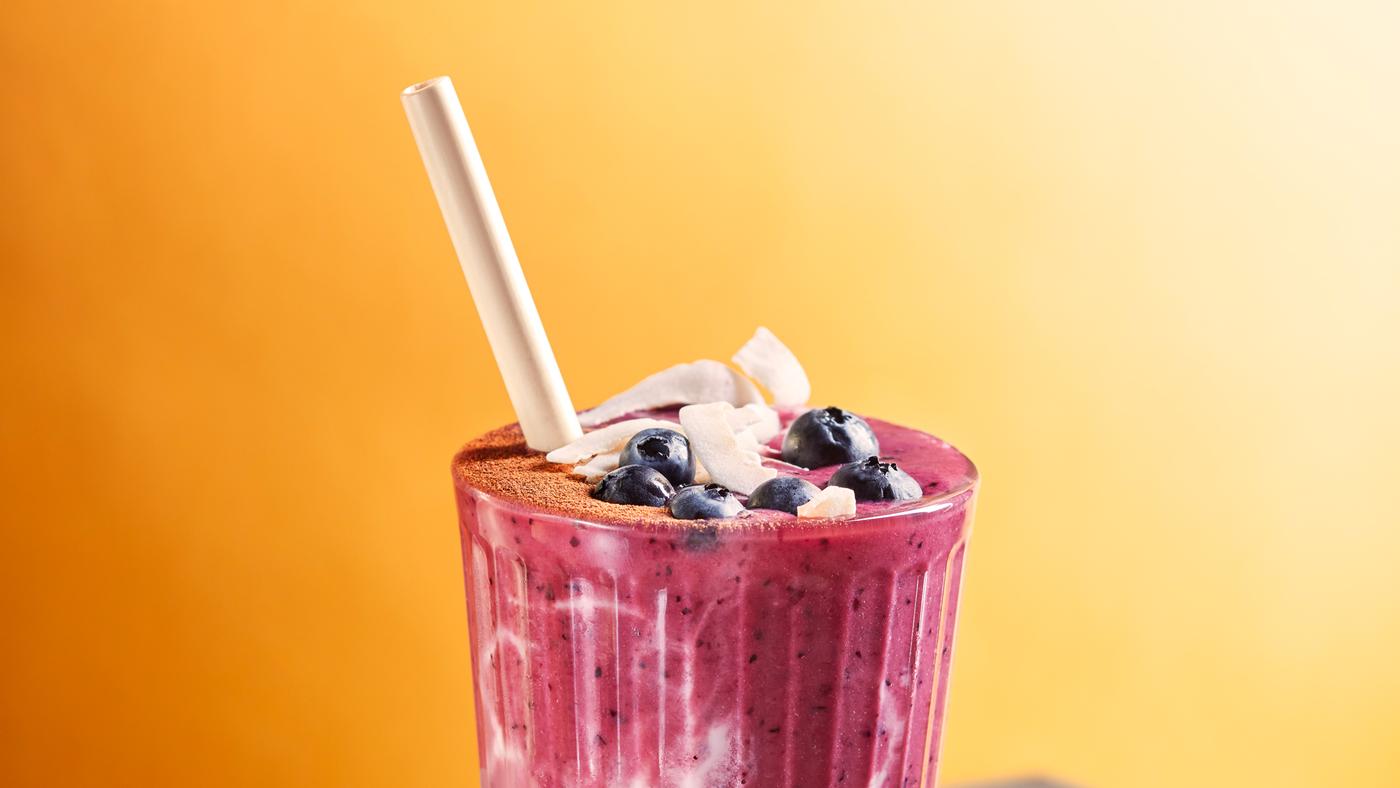 Pohár s ružovo-bielym smoothie z červenej repy a ovseného mlieka s bielou slamkou ochutené čučoriedkami a kokosovými plátkami.