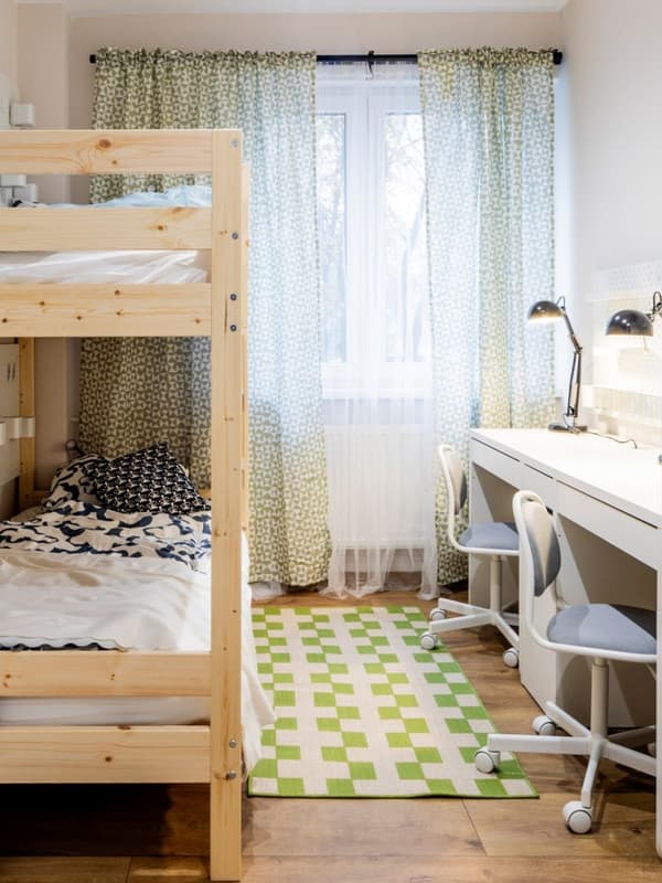 Pokój dziecięcy z drewnianym łóżkiem piętrowym po lewej stronie i dwom białymi biurkami po prawej. 