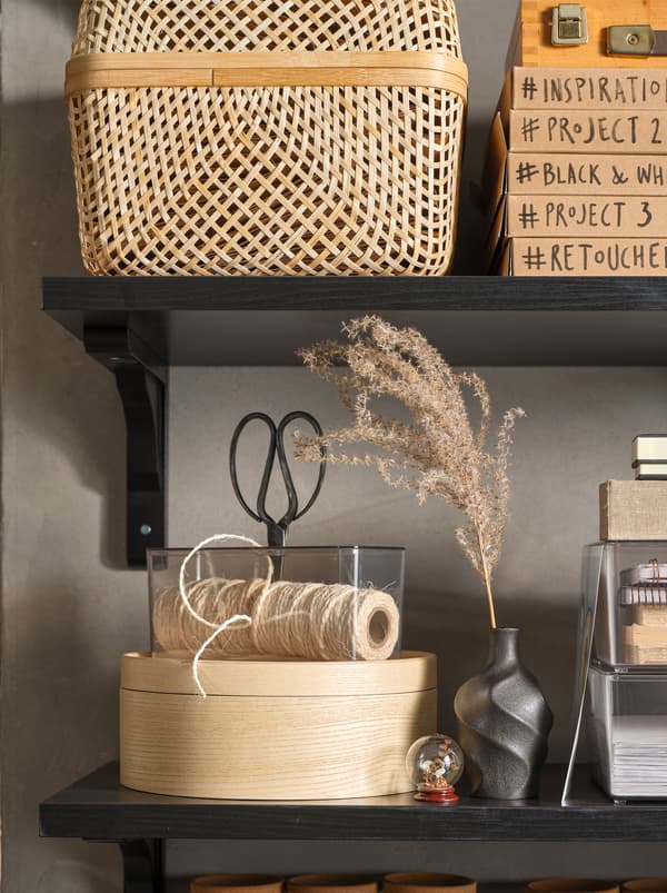 Polițe negre cu o cutie de depozitare SMARRA din bambus, o cutie rotundă MALLGRODA și obiecte personale pentru muncă și expunere.