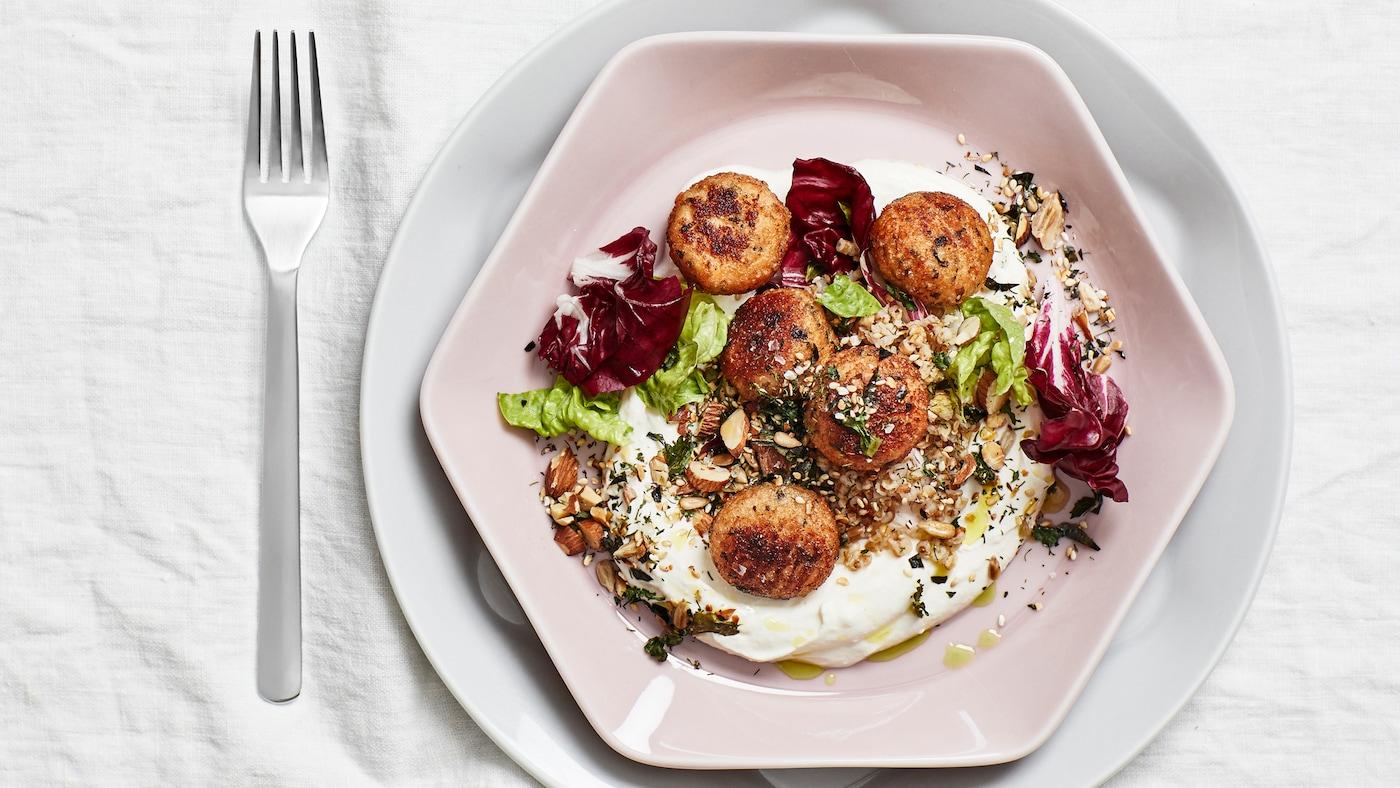 Polpette di salmone e merluzzo con mix di cereali, yogurt e furikake nordico su un piatto rosa con accanto una forchetta.