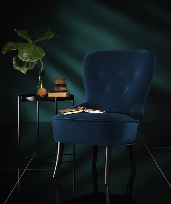 Poltrona in velluto blu scuro accanto a un tavolino in una stanza con pochi raggi di luce che penetrano.
