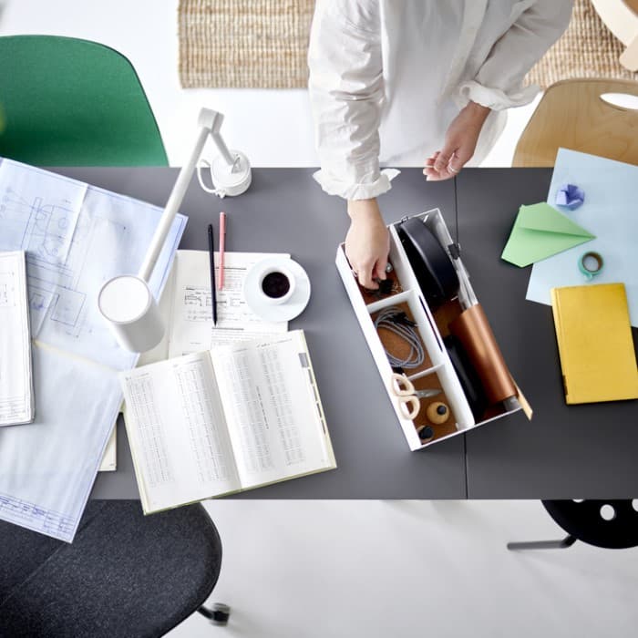 Poros rankų imamas daiktas iš baltos stalo dėžutės ant dviejų sujungtų stalų su darbo popieriais ir balta darbo lempa.