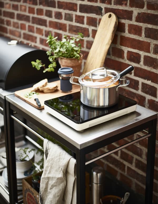 Portable induction hob with an IKEA PS 2017 pot and lid shown beside a barbecue on a terrace.