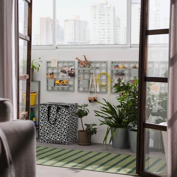 Porte aperte su un balcone con pannelli portaoggetti SNICKRA su una parete, una borsa nera e bianca, grandi piante in vaso e un tappeto - IKEA.