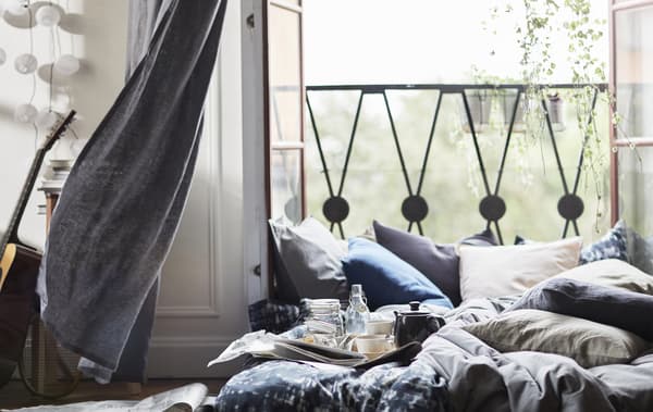 Portes-fenêtres ouvertes avec coussins et literie empilés sur un matelas posé à même le sol.