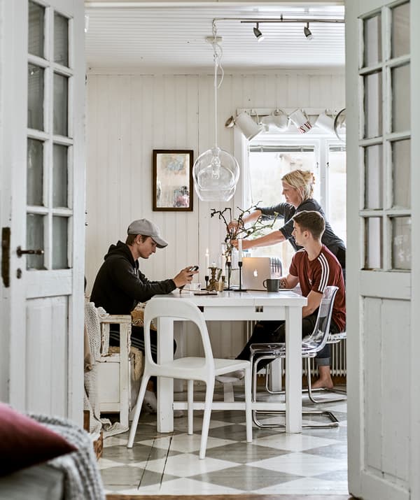 Portes-fenêtres ouvertes sur une salle à manger avec un sol en damier. Une famille est assise à une table blanche, des lampes en verre pendent au-dessus d’eux.