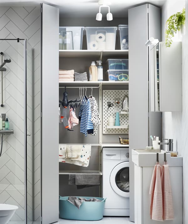 Portes grises entrouvertes cachant le coin buanderie, avec lave-linge, séchoir et boîtes sur des tablettes.