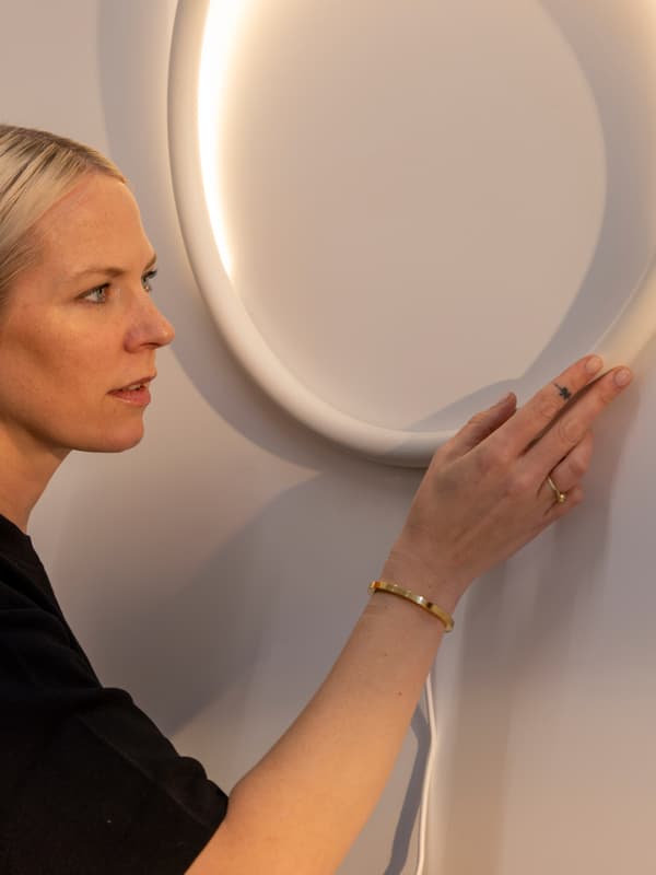 Portret oblikovalke Sabine Marcelis, oblečene v črno, ki pregleduje prototip VARMBLIXT okrogle stenske svetilke.