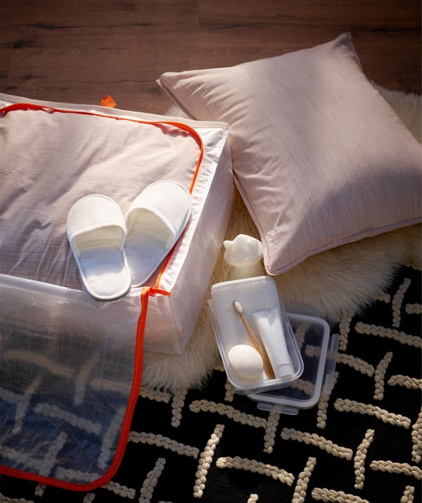 Posé sur le sol, un sac de rangement ouvert avec literie, pantoufles, brosse à dents et dentifrice, savon, serviette et veilleuse.