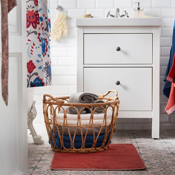 Posé sur le tapis de bain, le panier SNIDAD en rotin accueille des serviettes.