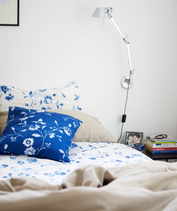 Posteľ s modro-bielymi bavlnenými posteľnými obliečkami s kvetinovým vzorom a lampou na čítanie v striebornej farbe na bielej stene nad ňou.