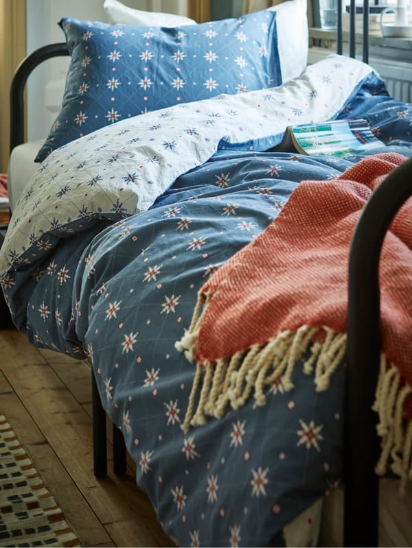 Posteľ s modrou posteľnou bielizňou ÅKERJORDFLY a oranžovou dekou ULLTISTEL.