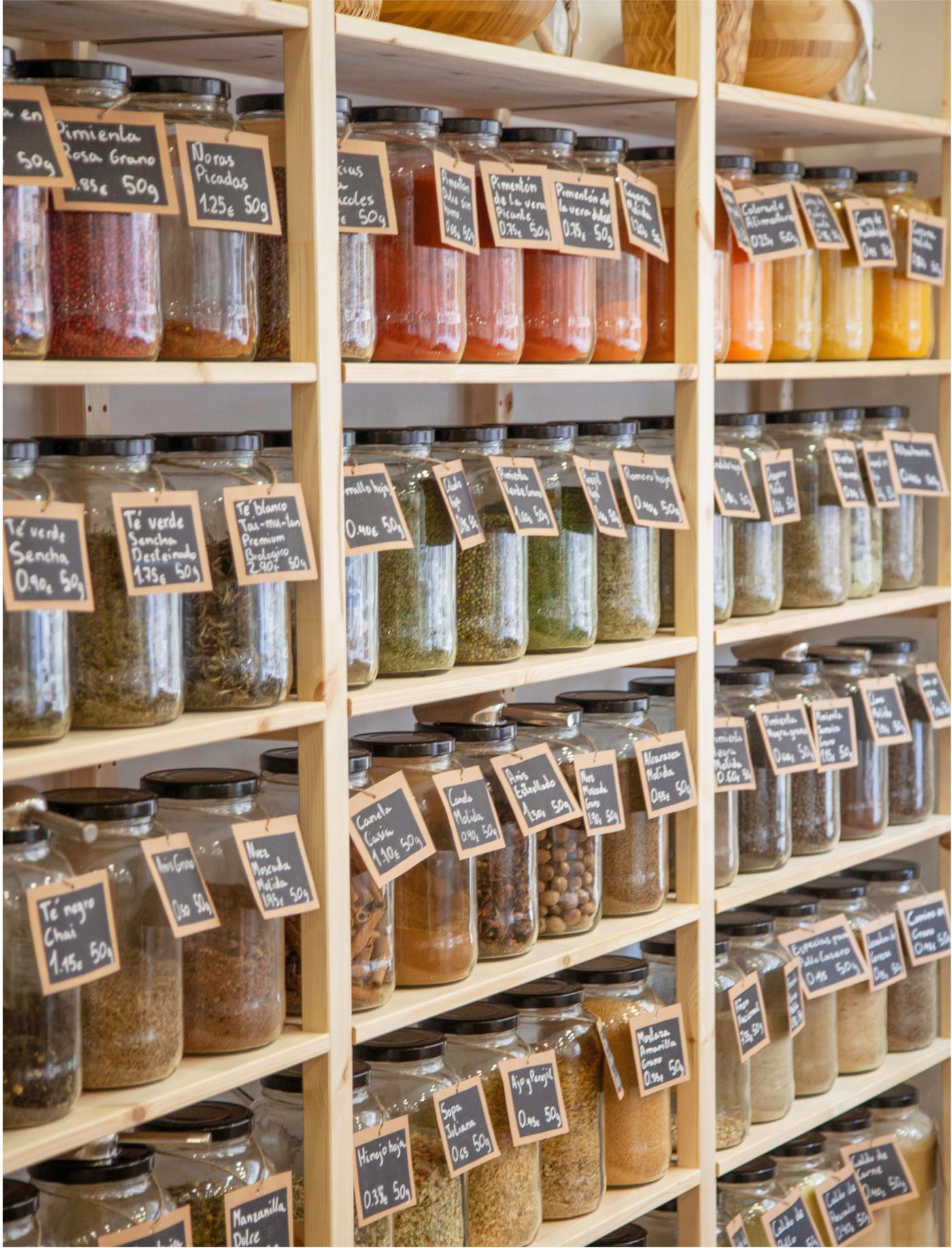 Pots de vidre amb espècies i herbes a prestatgeries de fusta, cada un amb etiqueta negra i text blanc.